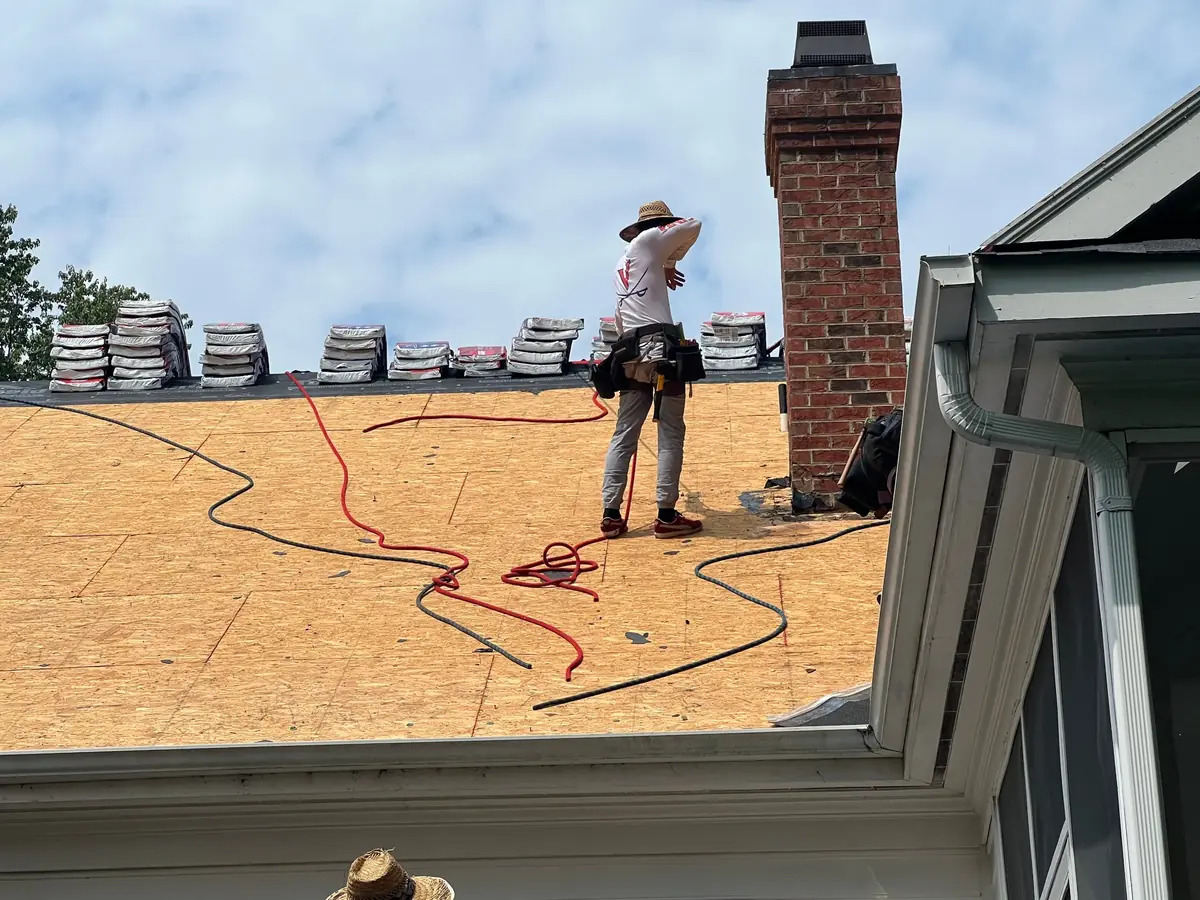 Roofer standing on bare OSB roof decking beside a brick chimney during a tear-off
