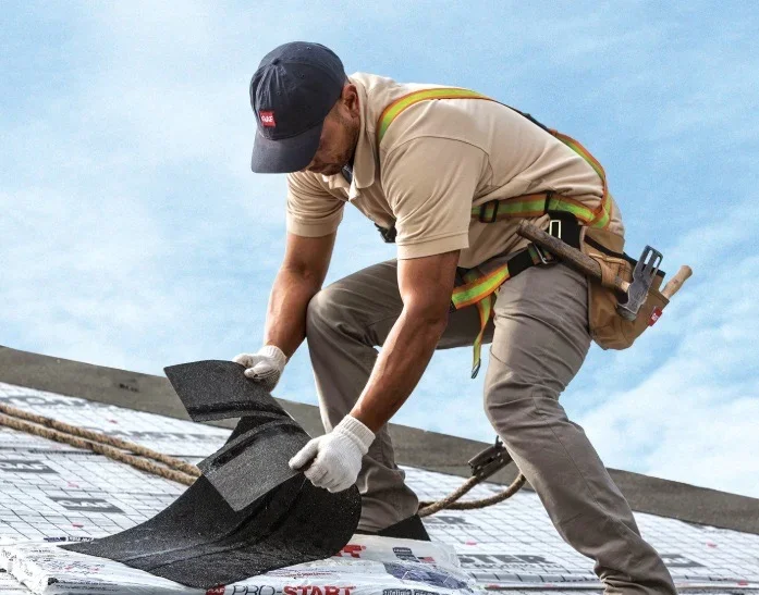 Roofer installing black asphalt shingles on residential roof with safety harness