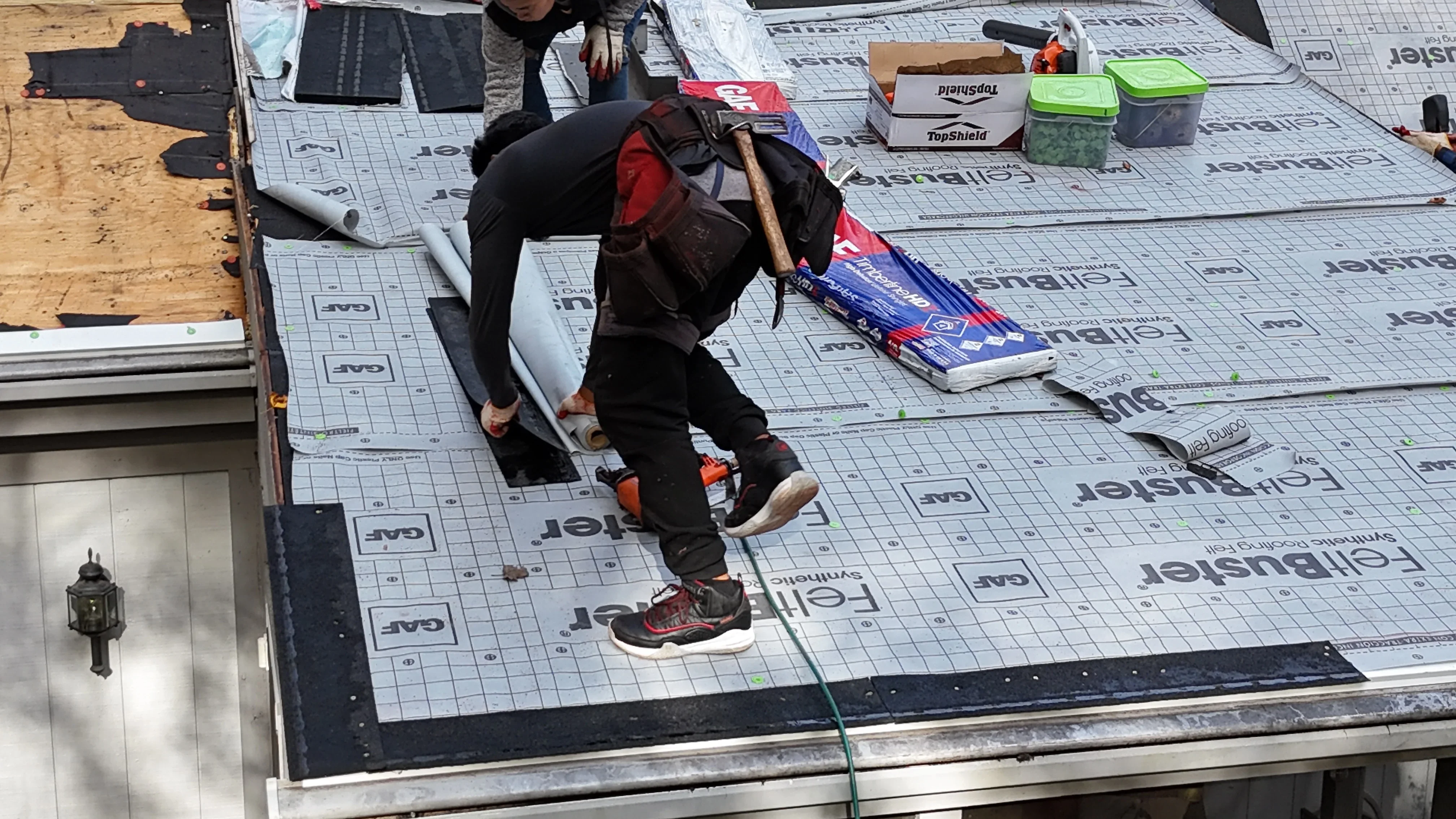Worker installing synthetic underlayment on plywood roof sheathing with grid pattern and fasteners