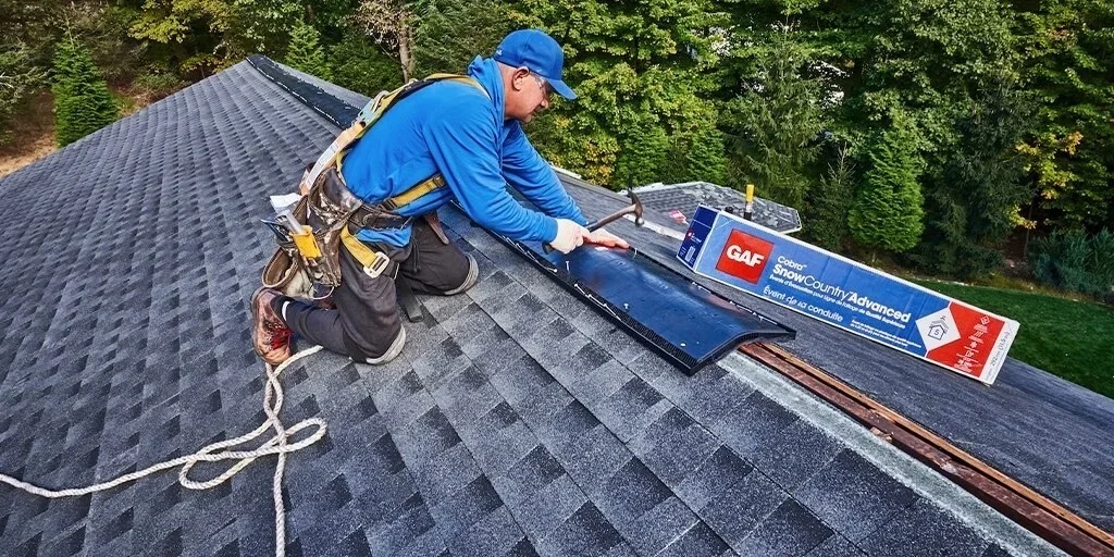 Roofer installing gray architectural shingles on residential roof with safety harness