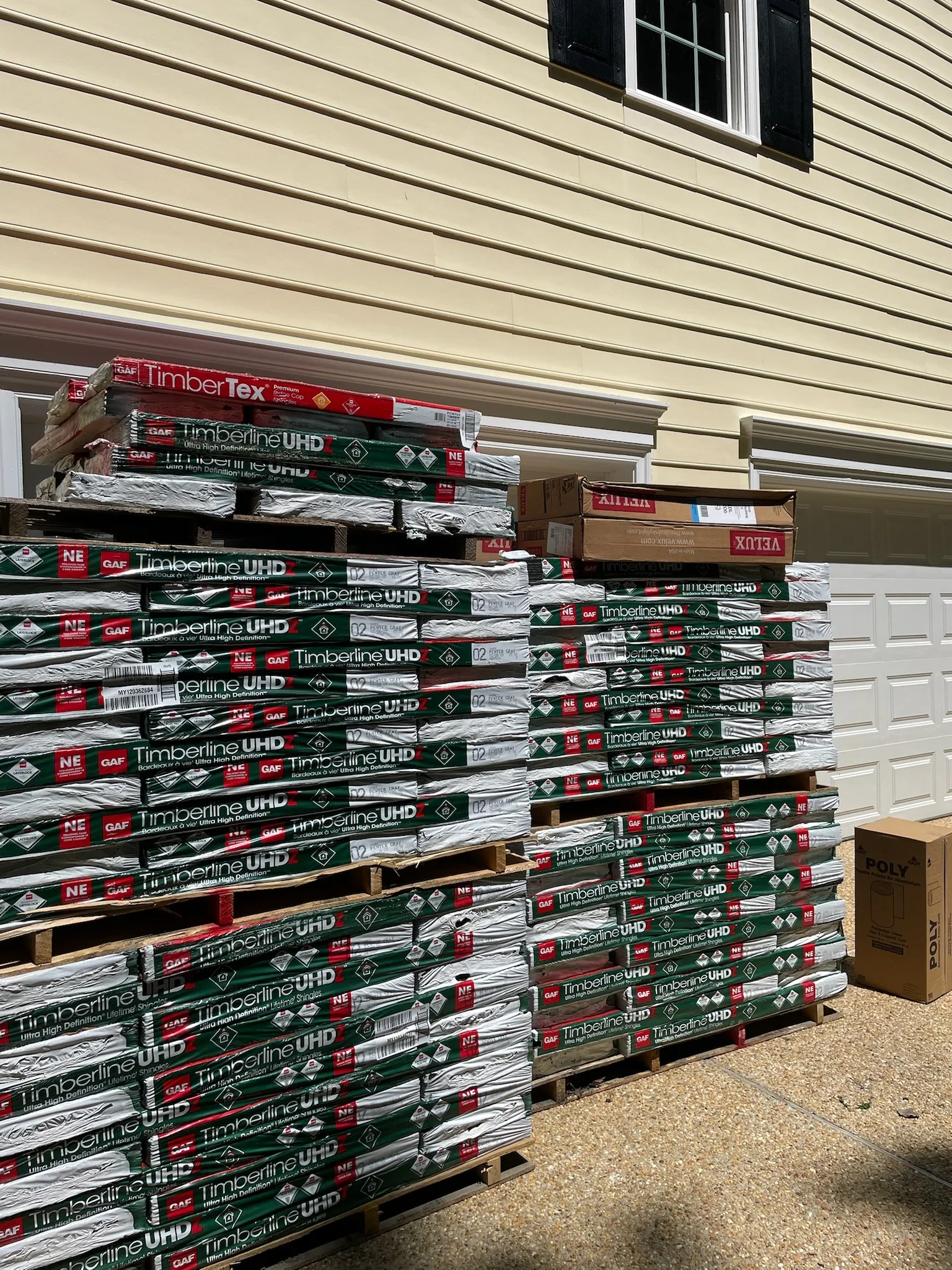 Stacked bundles of GAF Timberline UHD shingles and roofing materials on pallets against cream vinyl siding